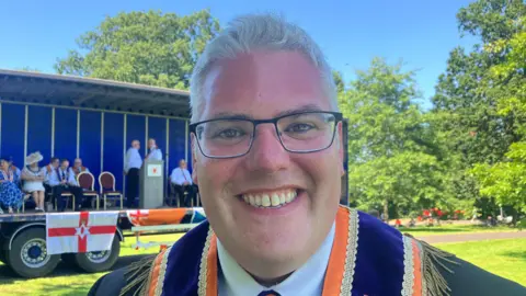 Thousands attending NI's Orange Order parades A man with short white hair and black rimmed glasses is smiling and looking into the camera. He is wearing a dark coloured suit jacket and an orange and purple sash.