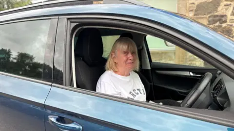 Airport drivers try to beat drop-off charges by parking in laybys Sheila McPheely sitting in the driver's seat of a blue car. She is smiling at the camera. She has blonde hair and is wearing a white T-shirt with grey writing on it.