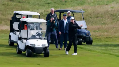 Security tightens as Donald Trump plays golf at Turnberry PA Media Donald Trump, wearing a blue jumper and trousers and a white USA cap, takes a golf swing on the fairway as other group members watch next to golf buggies