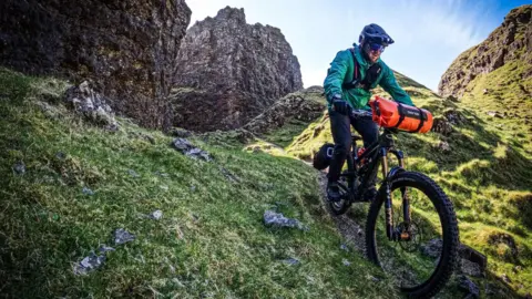 Mountain biker completes epic bid to ride down all 282 Munros Grizzly Munro Diaries A person on a mountain bike, wearing a black helmet and a green jacket, rides down a grassy hill.