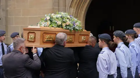 Funeral held for Norman Tebbit at Bury St Edmunds cathedral PA Media A light brown wooden coffin is carried on shoulders inside a doorway, young people in light blue shirts wearing berets stand guard either side of the coffin.
