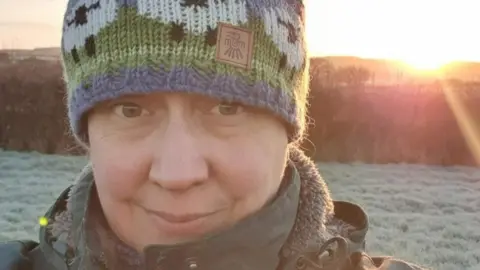 Electric vehicles to be discounted in government scheme Carolyn Hammond Carolyn Hammond smiling, wearing a knitted hat with a sunset and a field in the background.