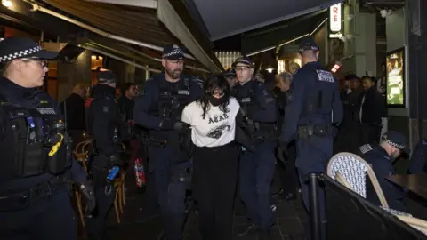Australian PM vows 'full force of law' after arson attack at Melbourne synagogue JOSH STANYER/EPA/Shutterstock Police take away an anti-Israeli protester with a black face mask, long dark hair and a white top from the restaurant where more police officers and others can be seen