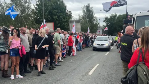 Hundreds of protesters gather at asylum hotel in Norwich area Richard Knights/BBC Scores of people standing on the road, many with St George's flags. A car is driving through the middle.