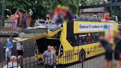 Fifteen hurt as double-decker bus hits bridge Salar Ardalani A number of people, who have been blurred out, are stood on the top floor of a double decker bus which has had its top sliced off. The back of the bus is handing off and people are watching on from either side of the road.