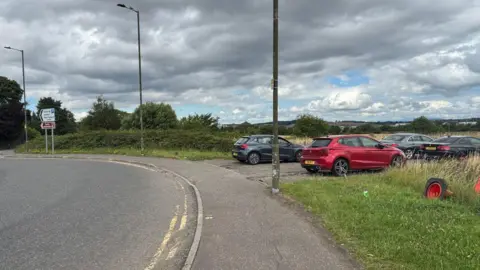 Airport drivers try to beat drop-off charges by parking in laybys A lay-by near a roundabout at Edinburgh Airport. There are four cars. The car closest to the camera is red. The cars further away are dark coloured, either black or grey.