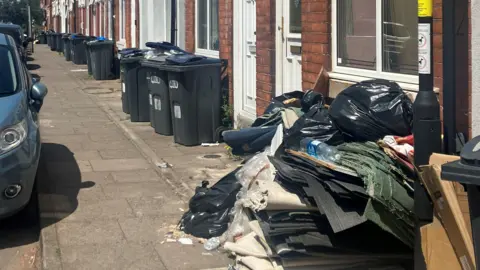 No end in sight for Birmingham bin strike after six months BBC Picture shows a terraced street with dark grey wheelie bins outside homes. In the foreground is a large pile of rubbish including black bags and strips of old carpet