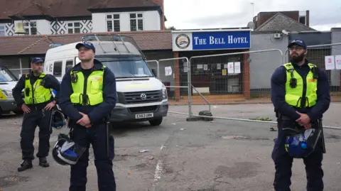 Epping council votes to urge government to shut asylum hotel PA Media Three police officers wearing hi-vis vests standing in front of vans and the hotel, which has metal fencing in front of it.