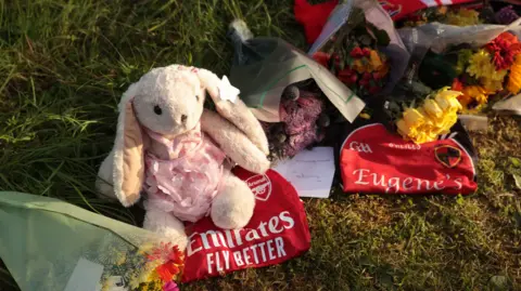 PA Media A rabbit teddy, flowers, an arsenal scarf and jersey and a GAA jersey are lying in grass 