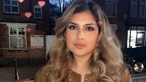 'Why I'm one of millions of UK adults not paying in' Saira Amir Saira Amir wearing gold earrings, Indian clothing, pink lipstick and looking at the camera. She is standing at street level, outside, with houses and one car behind her.