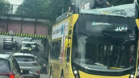 Fifteen hurt as double-decker bus hits bridge X People are standing on the top deck of a yellow bus of which the top has been pulled off. The front windscreen has been smashed.