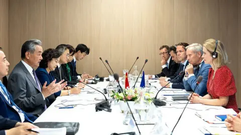 Getty Images Wang Yi and Kaja Kallas sitting at opposite ends of a table, facing each other. They are surrounded by Chinese and European officials.