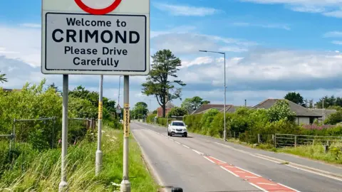 Woman who let child drive car in Crimond must do unpaid work A street sign that says welcome to Crimond beside a road with a lone white car under a blue sky with white clouds.