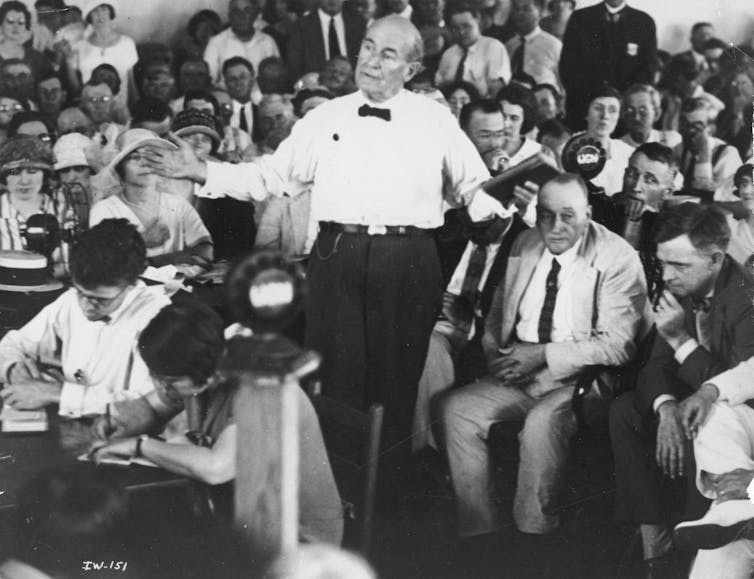 1 in 4 Americans reject evolution, a century after the Scopes monkey trial spotlighted the clash between science and religion A man in a white shirt and black bow tie stands with arms outstretched while talking, while others seated in rows around him look on.