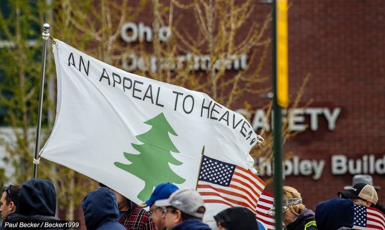 A white flag bearing the words 'An Appeal to Heaven,' featuring a green pine tree, with the American flag displayed beneath it.