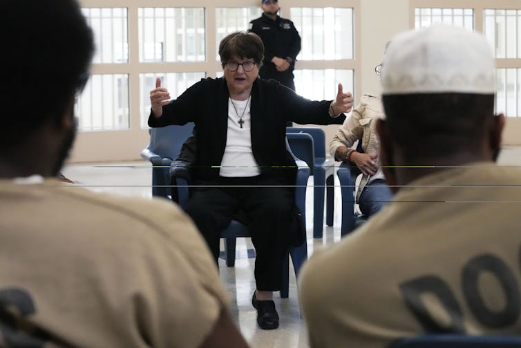 A seated woman in a black blazer and a cross necklace gestures as she speaks with other people seated in a circle.