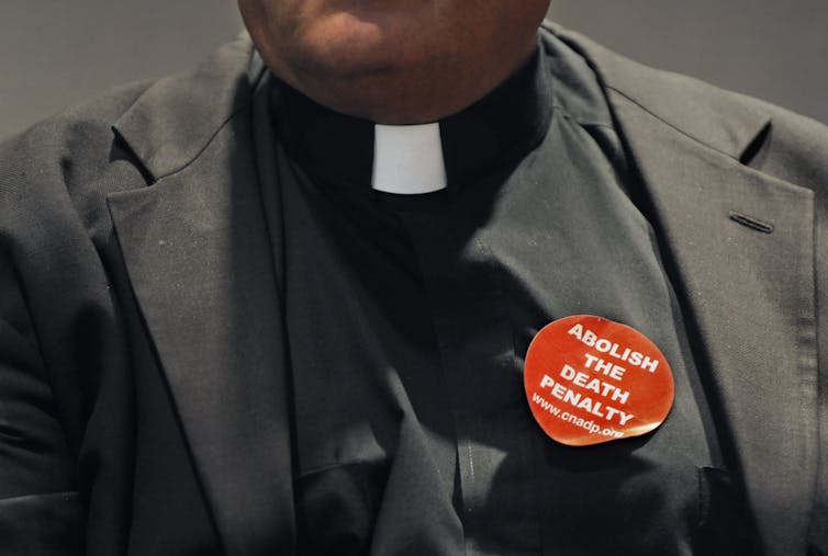 A close-up photo of the chest of a man wearing a priest's collar and a red sticker that says 'abolish the death penalty.'