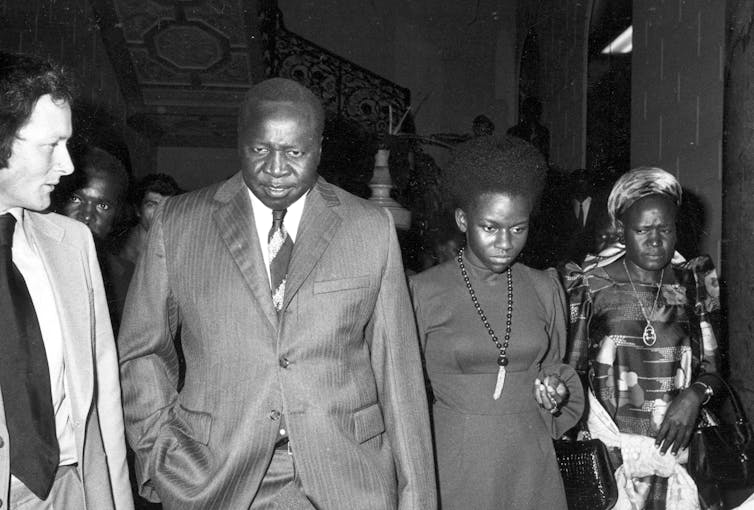 A Black man in a suit walking with a woman and another dressed in traditional African attire.