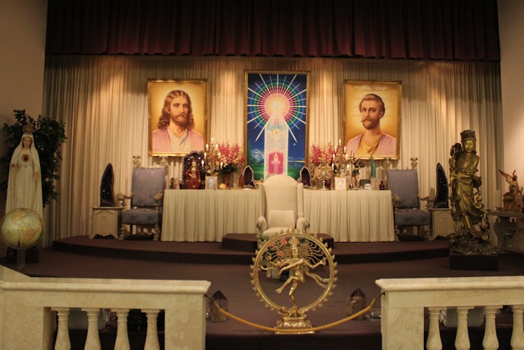 A stage area displaying an image of the Hindu god Shiva and two large portraits of men, with a white chair flanked by two flower-laden tables.
