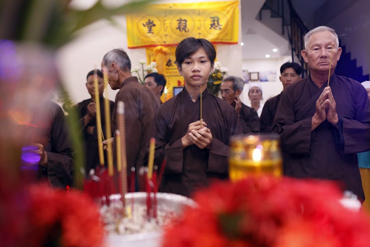 It is becoming easier to create AI avatars of the deceased − here's why Buddhism would caution against it A young man, holding incense sticks, stands with an elderly person while they both fold their hands in prayer at an altar, with several others behind them.