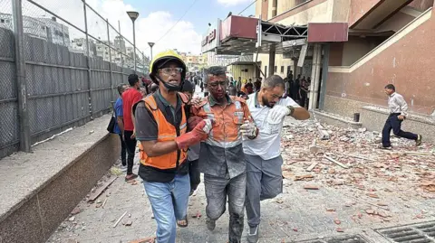 At least eight killed in Israeli strike on Gaza hospital, health ministry says Getty Images An injured emergency worker is led away by two other emergency workers. There is rubble and wreckage behind them. He is badly hurt.
