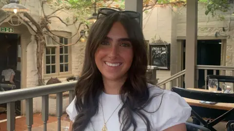 Working class students divided on civil service summer internship reform A photo of Nell Ashworth smiling as she sits at a restaurant. She is wearing a white blouse with a gold pendant and has long brown hair.