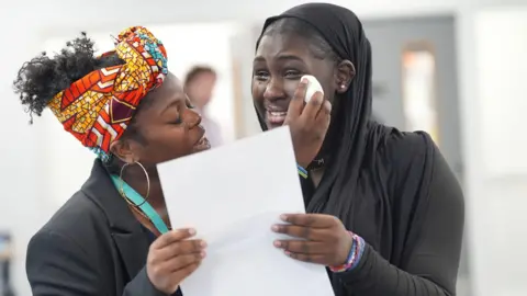 PA Media A young woman (l) wearing a black headscarf cries happy tears as she shows her GCSE results to her teacher, a woman wearing a dark suit, a colourful headwrap and large hoop earrings. 