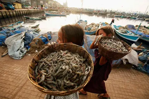 Inside the Indian factories hit hard by US's 50% tariffs Bloomberg via Getty Images Two young girls carry shrimps in baskets on a jetty in Ratnagiri, India
