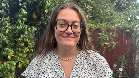 Under-22s join call for free travel Gracie Moore A young woman wearing square, tortoiseshell glasses and a white and blue floral shirt smiles while standing in front of a tree laden with pears and a red wooden fence covered in vines.