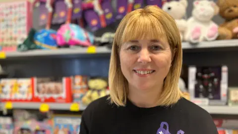 Southampton social supermarket alternative to food banks Becky is looking into the camera. She has blonde hair in a shoulder-length bob. She's wearing a black T-shirt with a purple shopping bag logo, and in the background toys and board games can be seen slightly out of focus.