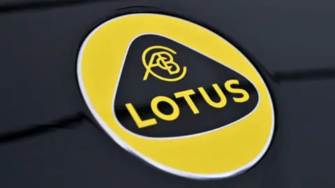Sports car manufacturer Lotus to cut 550 jobs at Norfolk base Getty Images The Lotus logo. It is a yellow circle which says LOTUS in yellow writing. The writing sits inside a curved triangle.