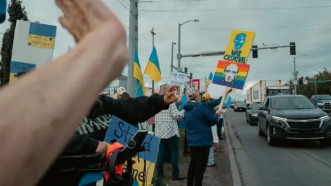 Alaskans share anger and hope as Trump and Putin fly in People holds signs beside a road one saying 'war criminal'