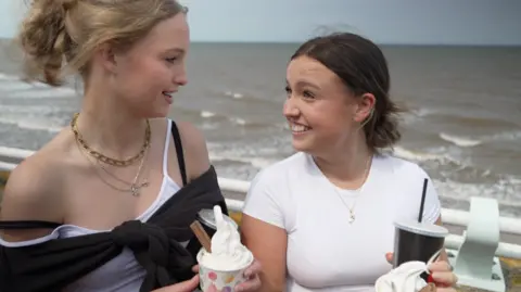 Solar-powered ice cream maker has a record sunny year in Somerset BBC Two teenage girls turn to smile at one another. They are both holding Mr Whippy-style ice creams with chocolate flakes, in tubs. The sea is not far behind them.