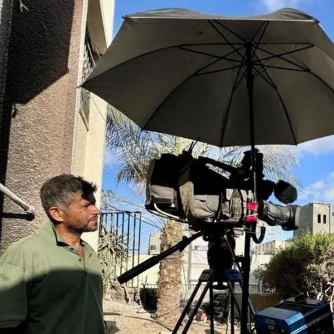Israeli double strike on Gaza hospital Reuters A man stands to the side of a TV camera watching it as he films. He is stood under an umbrella in Gaza