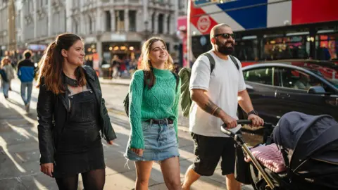 UK government borrowing lower than expected in July Getty Images Two women and a man who is pushing a buggy with a little girl in it along a street in London with a bus in the background