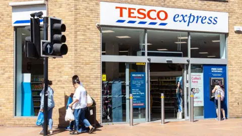 Tesco meal deal price rises to £3.85 for loyalty card holders Getty Images Three people walk past a Tesco Express carrying plastic shopping bags, while someone else uses a Tesco ATM at the front of the store.