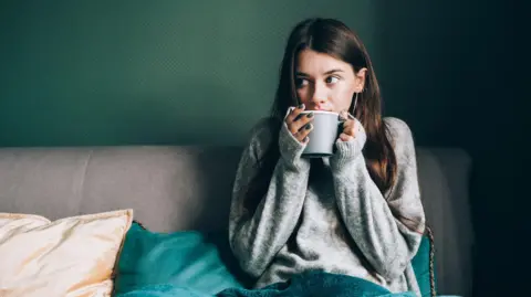 Four ways to keep your energy bills down Getty Images Stock photo shows a woman in a jumper under a blanket with a mug of tea. She is hunching her shoulders as if feeling cold while sitting on the sofa at home.