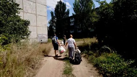 Inside Donetsk as residents flee attacks on Ukrainian region Putin wants to control Three people walk down a dirt path past a building and piles of weeds, carrying large bags