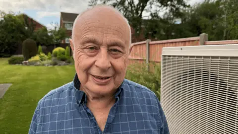 Tamworth cafe owner feared £24k E.On energy bill would sink firm Dr David Toke standing in a garden. There is a heat pump next to him. He is balding with white hair and he is smiling. He is wearing a blue checked shirt which is open at the neck.