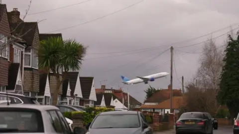 Heathrow Airport's expansion plans to cost £49bn, plans reveal A row of suburban houses with a plane taking off very close to the houses. There are cars in the road in front of the houses.