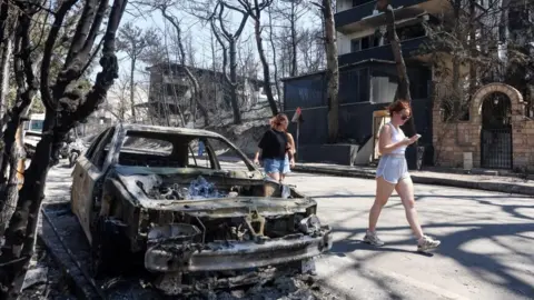 Wildfires rage across southern Europe as temperatures pass 40C Reuters Women in shorts walk past a fully charred and burnt car ion a street surrounded by charred trees