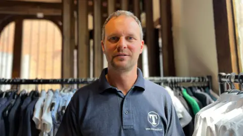 School uniform costs 'extortionate' say parents in Hull Becki Bowden/BBC A man with short grey hair and wearing a dark coloured polo shirt stands in a hall surrounded by clothes rails.