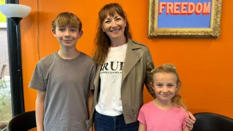 How to feed children for less in the holidays holidays Coletta Todd with long ginger hair and a fringe standing between her 14-year-old son henry who is wearing a grey t shirt and her sweven-year-old daughter Mary who is wearing a pink t-shirt. They are standing in front of a bright roange wall with a lamp to one side and gold frame on the wall with the words Freedom in typographical art