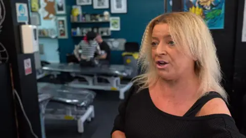 Covid loss businesses take action against insurer BBC A middle-aged woman with shoulder-length light blonde hair. She wears a black shirt and stands in a tattoo studio with several tattoo beds behind her.