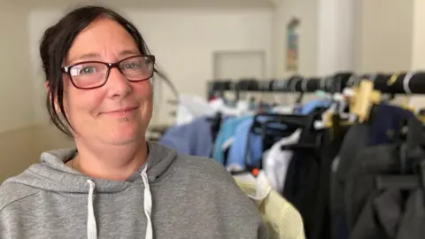 Forest of Dean school uniform pop-up a 'godsend', mum says BBC A woman with tied-up dark hair, glasses and a piercing on her bottom lip and chin smiles as she stands in front of a clothing rack full of school uniforms. She is wearing a grey hoodie with white strings.