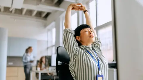 Perfect posture is a myth Getty Images A busy woman takes a moment to stretch while working at her desk in a modern office, balancing productivity and well-being