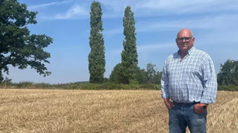 Herefordshire farmer sees broccoli harvest hit by heatwave and lack of water BBC/Malcolm Prior Martin, dressed in a light-blue-checked shirt and blue jeans, stands with his hands in his pockets in a wheat field that has been harvested. He is bald and wearing rectangular glasses. A blue sky with little cloud indicates it is still hot while the yellow crop field appears dry. On the horizon are green trees.