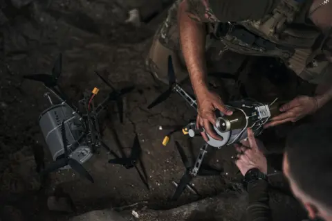 They escaped Ukraine's frontlines. The sound of drones followed them Getty Images An arm holds on to a silver-coloured drone perched on the ground in the dark