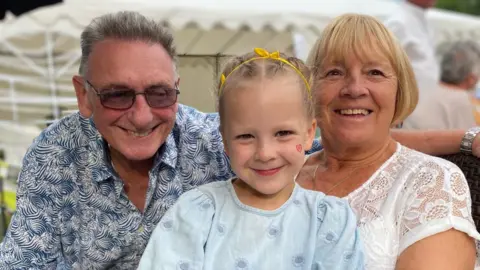 Holidays with grandparents 'on the up' amid summer childcare cost Avril Hackett and her husband with their granddaughter Emily in between them. She is holding a colourful teddy and there is a large, white marquee-style tent behind them