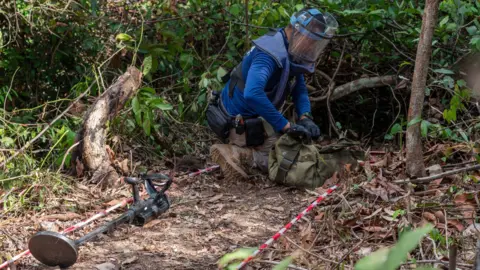 A fierce war of words keeps the two countries on edge BBC/ Jonathan Head A Thai soldier wearing blue protective gear and a blue helmet demonstrates how they detect and handle mines - but it's not a real one on site. One of the Thai soldiers was injured by a PMN-2 less than 100 meters from there on 9 August.